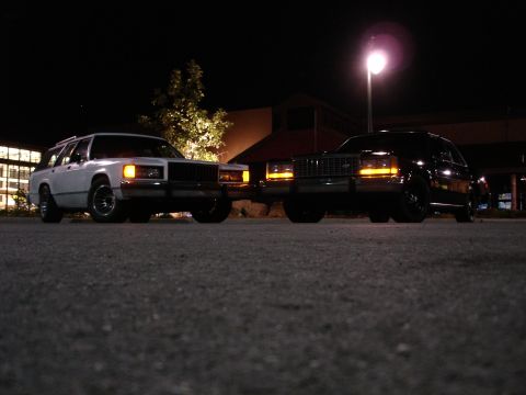 1987 Ford LTD Crown Victoria - Low ground-level night photo of two full-size cars parked nose-to-nose in a lot with parking lamps lit: a white station wagon with roof rac…