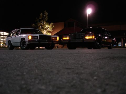 1987 Ford LTD Crown Victoria - Two station wagons are parked side-by-side at night. The one on the left is white, and the one on the right is black. Both vehicles have th…