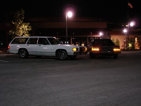 1987 Ford LTD Crown Victoria - Two station wagons are parked in a parking lot at night. The white station wagon is on the left and a dark-colored station wagon is on the…