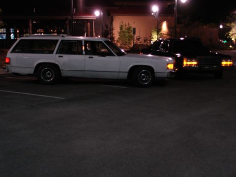 1987 Ford LTD Crown Victoria - A white station wagon is parked next to a black sedan at night in a parking lot. The station wagon is a Ford Crown Victoria, and the sedan…