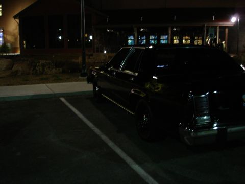 1987 Ford LTD Crown Victoria - The rear passenger side of a black sedan is parked in a dimly lit parking lot at night. The vehicle is parked next to a curb with a white p…
