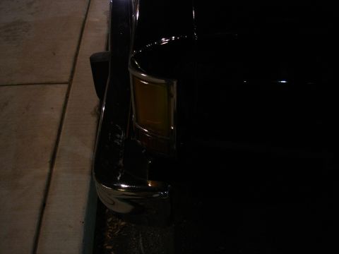 1987 Ford LTD Crown Victoria - Dark night close-up of the front corner of a black car parked along a concrete sidewalk, showing the amber parking/turn signal lens set in…