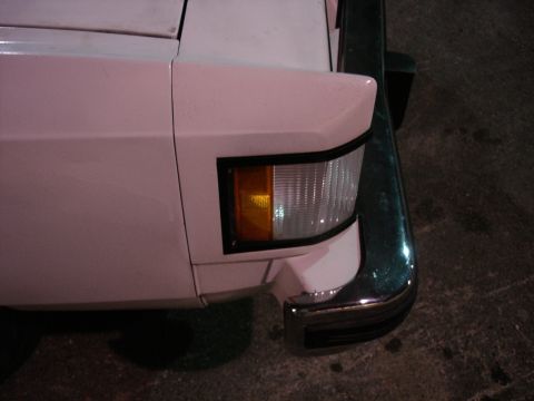 1987 Ford LTD Crown Victoria - Night close-up of the front right corner of a white car, showing the wraparound amber-and-clear side marker/turn lamp set flush in the fend…