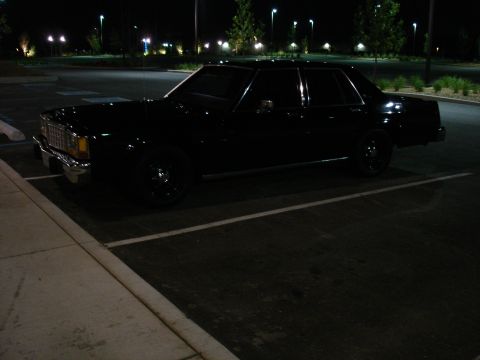 1987 Ford LTD Crown Victoria - A black sedan parked in a dimly lit parking lot at night. The vehicle is viewed from the front quarter angle, showing its chrome grille and…