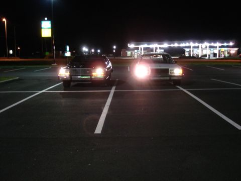 1987 Ford LTD Crown Victoria - Two sedans are parked in a parking lot at night, with their headlights on. A gas station with bright lights is visible in the background.