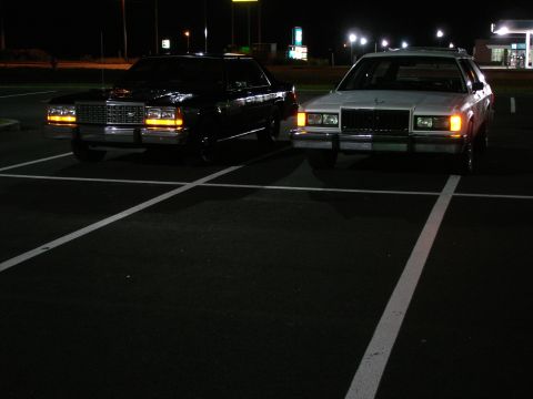 1987 Ford LTD Crown Victoria - Two full-size sedans are parked in a dark parking lot at night, with their headlights illuminating the asphalt. The car on the left is blac…