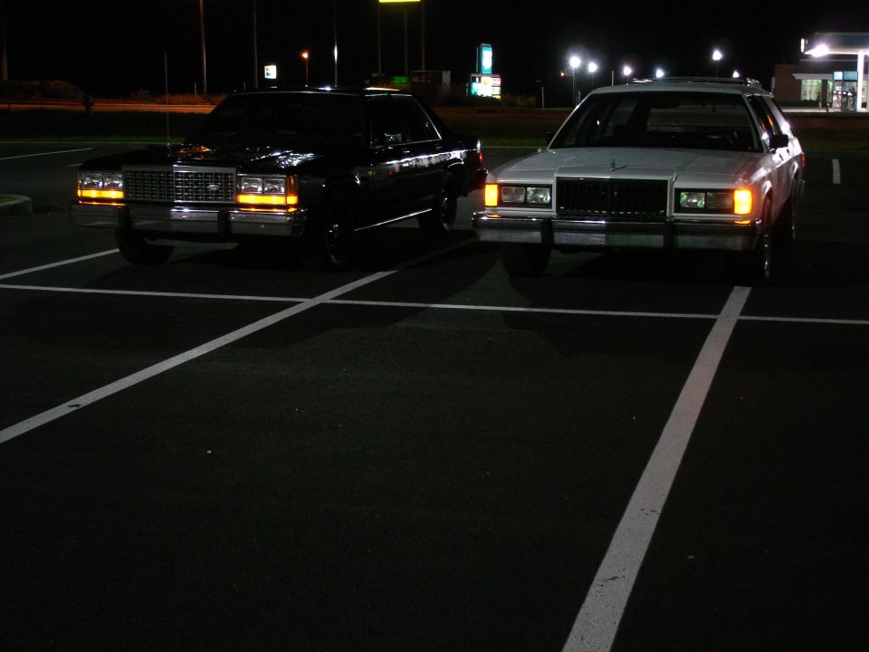 Two full-size sedans, one black and one white, parked in a dark parking lot at night.
