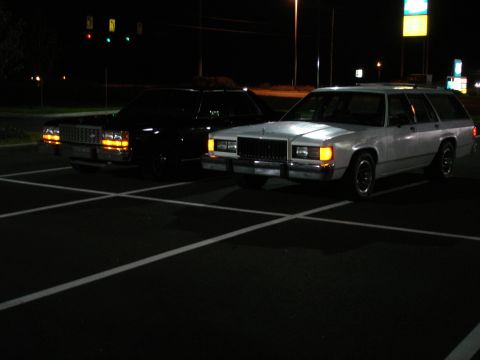 1987 Ford LTD Crown Victoria - Two station wagons are parked in a dark parking lot at night. The car on the left is black, and the car on the right is white.