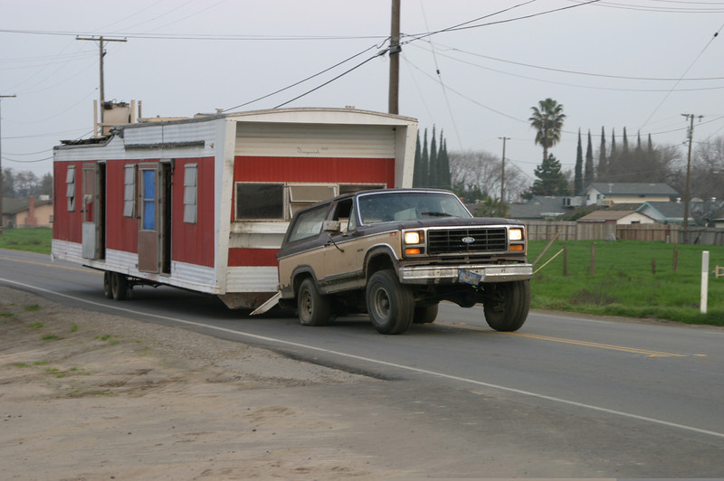 Brown two-tone Ford Bronco towing a red and white mobile home on a rural road.