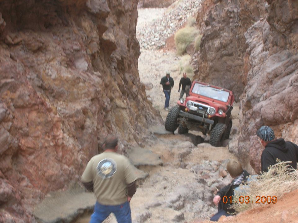 Red classic SUV driving down a rocky canyon with people watching.
