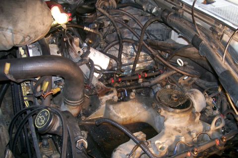 1992 Ford C-350 (Centurion Conversions) - A close-up view of a dirty diesel engine bay, showing various hoses, wires, and engine components. A bright light illuminates some of the p…