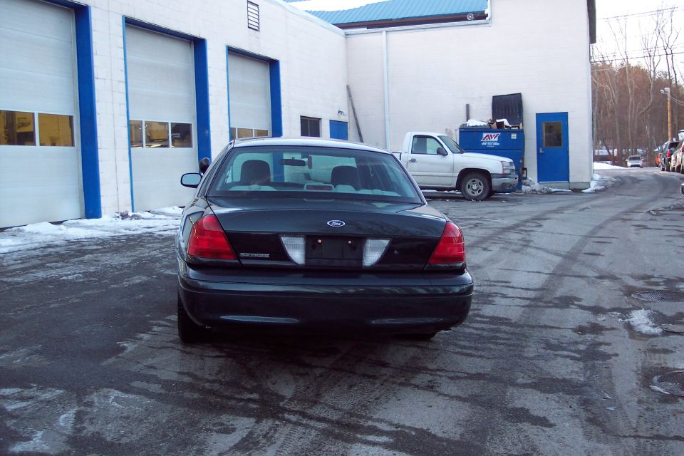 Rear view of a dark Ford Crown Victoria parked in front of a garage with a white pickup truck nearby
