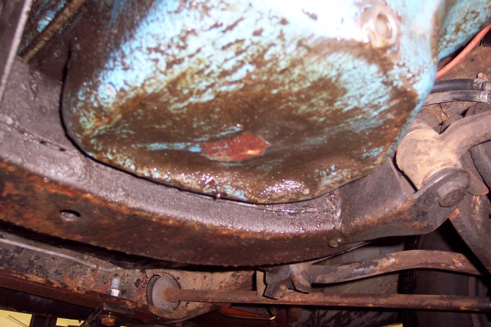 Underside of a vehicle showing a grimy oil pan and rusted frame.