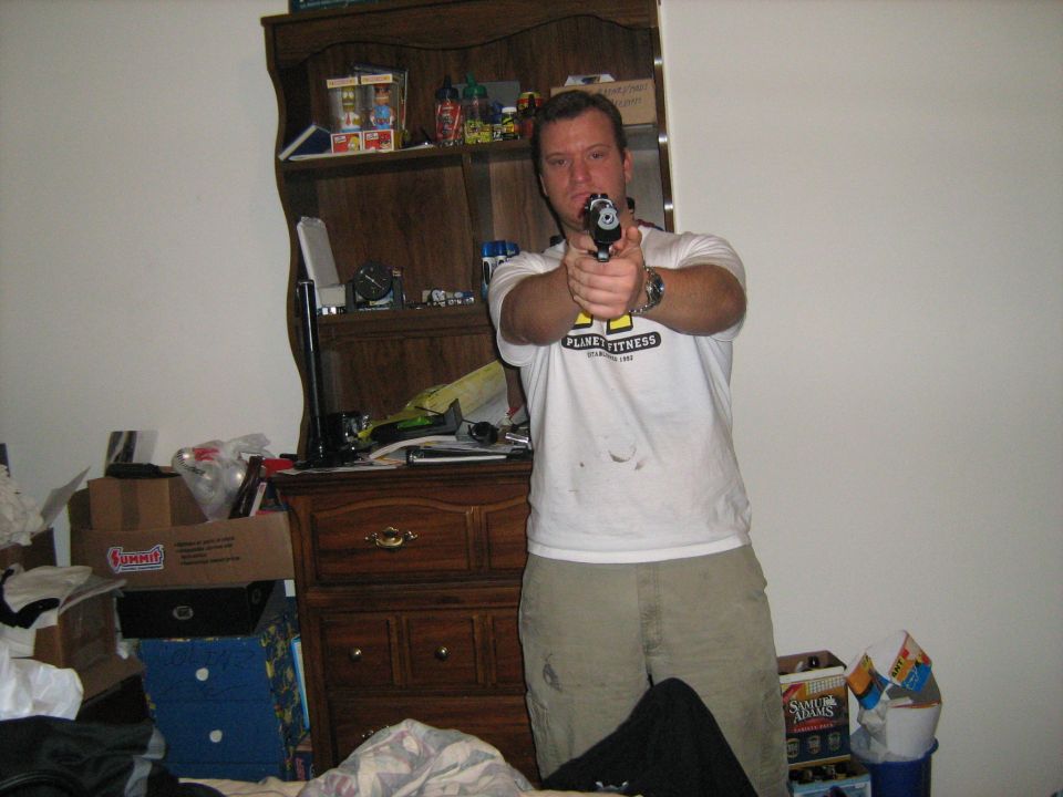 Man in a white t-shirt pointing a handgun toward the camera in a cluttered bedroom