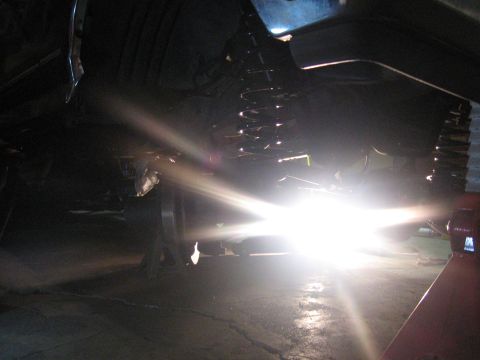 1989 Ford Bronco - The undercarriage of a vehicle is visible, supported by jack stands, with a bright light creating lens flare.