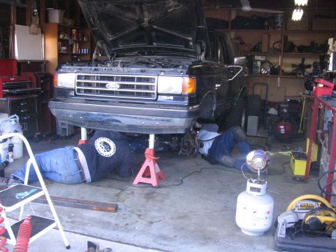 1989 Ford Bronco - Two mechanics work on a black 1989 Ford Bronco in a cluttered garage, with the vehicle raised on jack stands and its hood open.