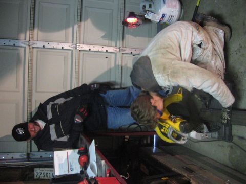 1989 Ford Bronco - Two individuals are engaged in work with a chop saw, set against a garage backdrop with a propane tank and overhead door.