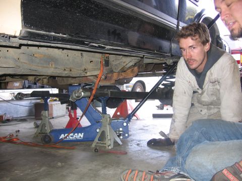 1989 Ford Bronco - Mechanics work on the undercarriage of a vehicle supported by jack stands in a garage.