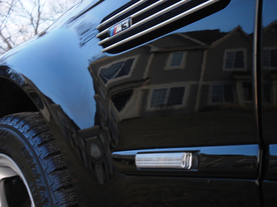 Black car fender with M3 badge and side marker light, reflecting a house