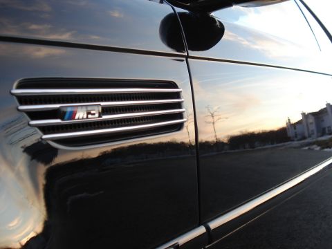 1998 Ford Taurus - Close-up of a black car's fender, highlighting the M3 badge and chrome trim, with reflections of the sky and buildings.