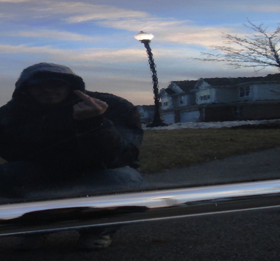 Reflection of person in hooded jacket in shiny car door, with houses and streetlamp in background