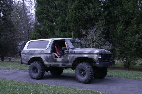 1979 Ford Bronco - This lifted Ford Bronco, painted in camouflage, sits on a paved surface with trees in the background.