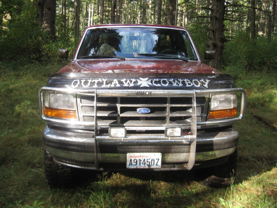 Lifted red Ford pickup with brush guard and OUTLAW COWBOY hood lettering in woods
