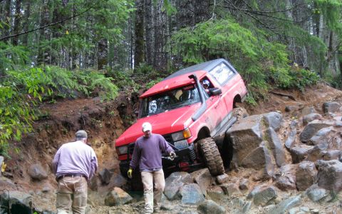 1989 Ford Bronco - A red SUV with a black top is shown off-roading on a steep, rocky incline.