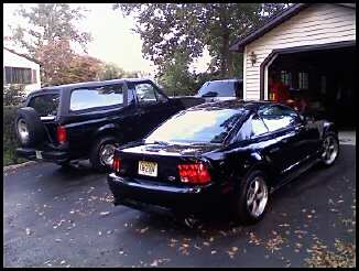 1996 Ford Bronco - Two black vehicles, a Ford Bronco and a Ford Mustang, are parked in a driveway next to a garage.