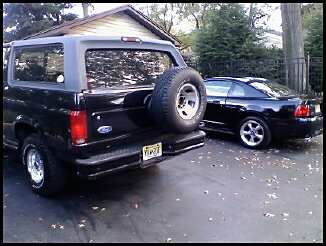 1996 Ford Bronco - A black 1996 Ford Bronco is seen from the rear, with a spare tire on the tailgate. A second black car is parked to the right.
