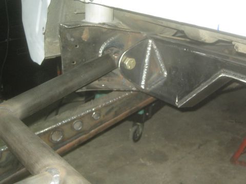 1990 Ford Bronco - close-up of a welded steel suspension link bolted to a gusseted bracket on a bare metal chassis in a shop.