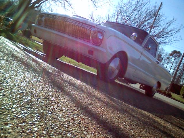 Low-angle front-left view of a light silver vintage Ford with chrome grille, parked by a gravel lot.