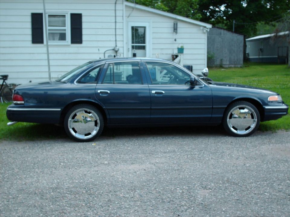Dark blue Ford Crown Victoria sedan on chrome wheels parked beside a white house