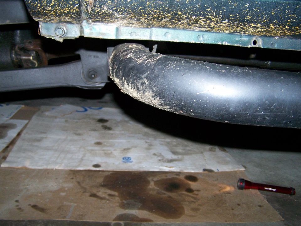 Underside of a vehicle with exhaust pipe, suspension, oil stains, and a flashlight.