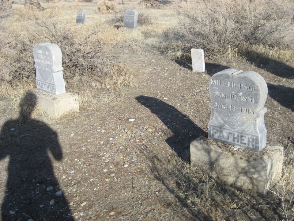Cemetery scene with headstones and dry brush, person's shadow in foreground.