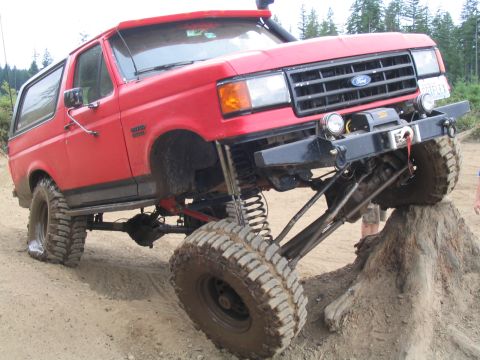 1989 Ford Bronco - A bright red, lifted Ford Bronco is shown at an extreme angle, with its front tire resting on a dirt mound. The vehicle features large off-…