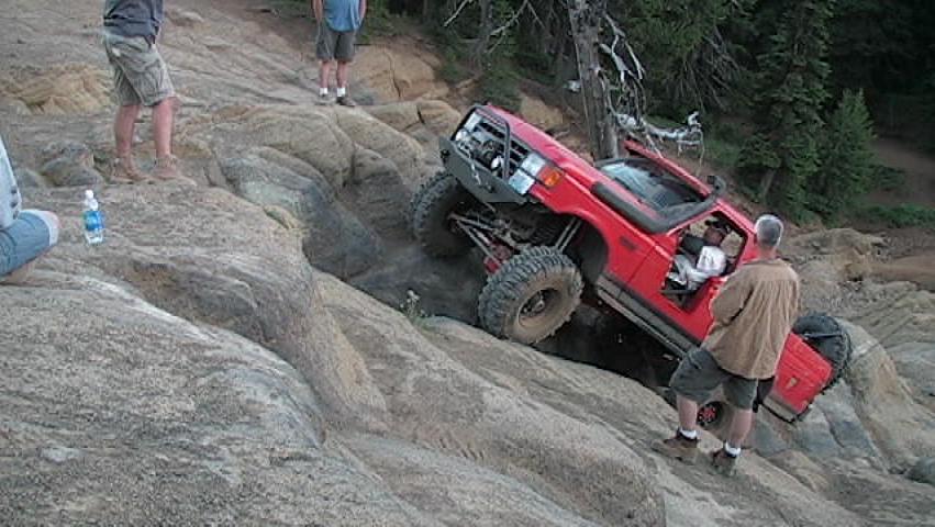 Red Ford Bronco climbing a steep rocky hill with people watching.
