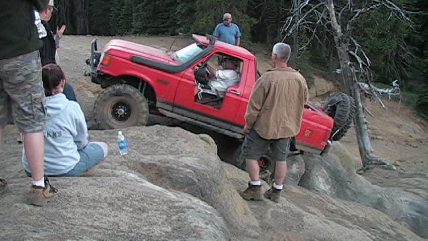 Red Ford Bronco off-roading over rocks with people watching.