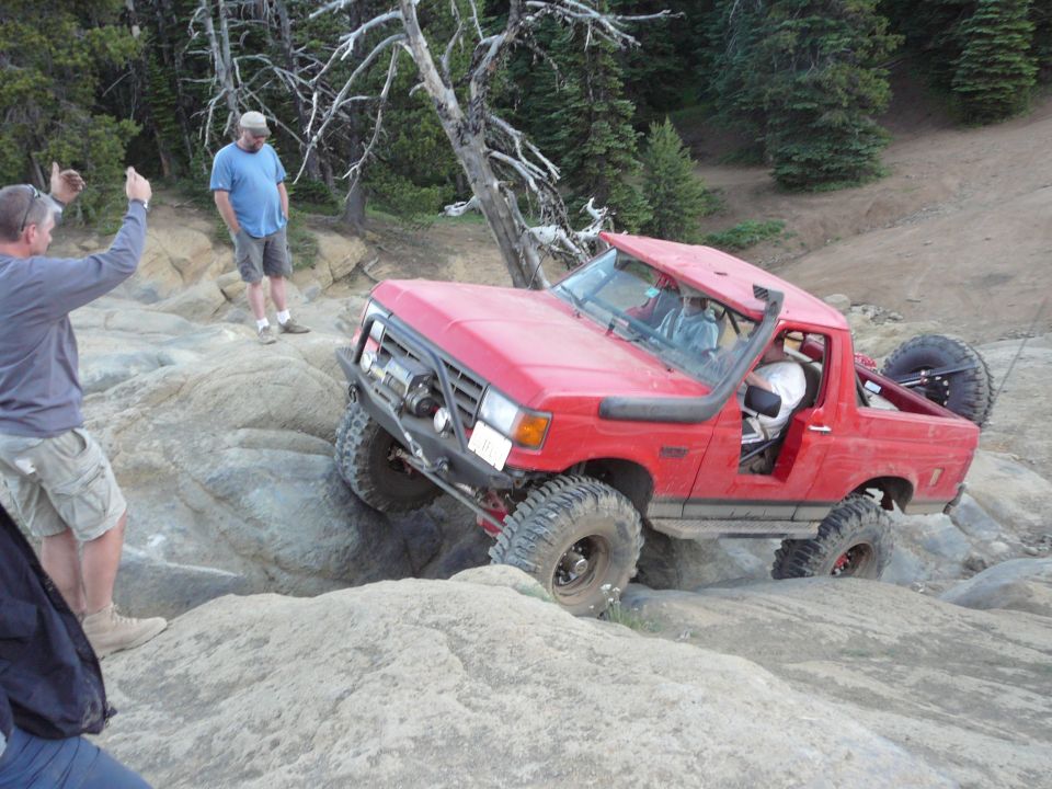 Red Ford Bronco off-roading on a rocky trail with two men watching.