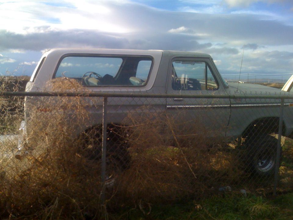 Gray 1983 Ford Bronco side view behind a fence with dry brush and cloudy sky.