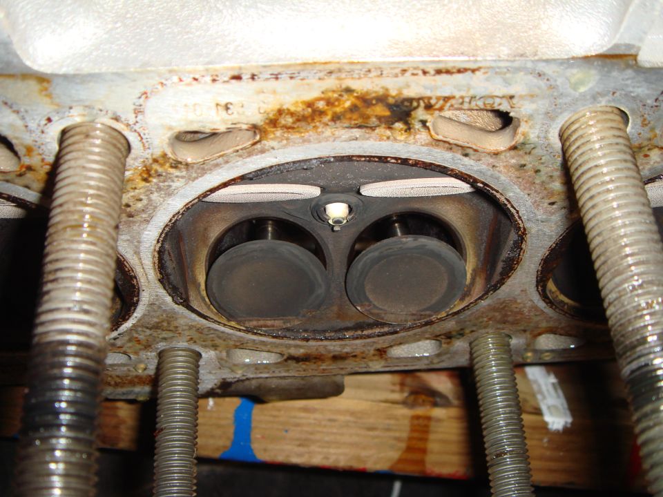 Close-up of rusty cylinder head combustion chamber with two valves and spark plug, flanked by studs.