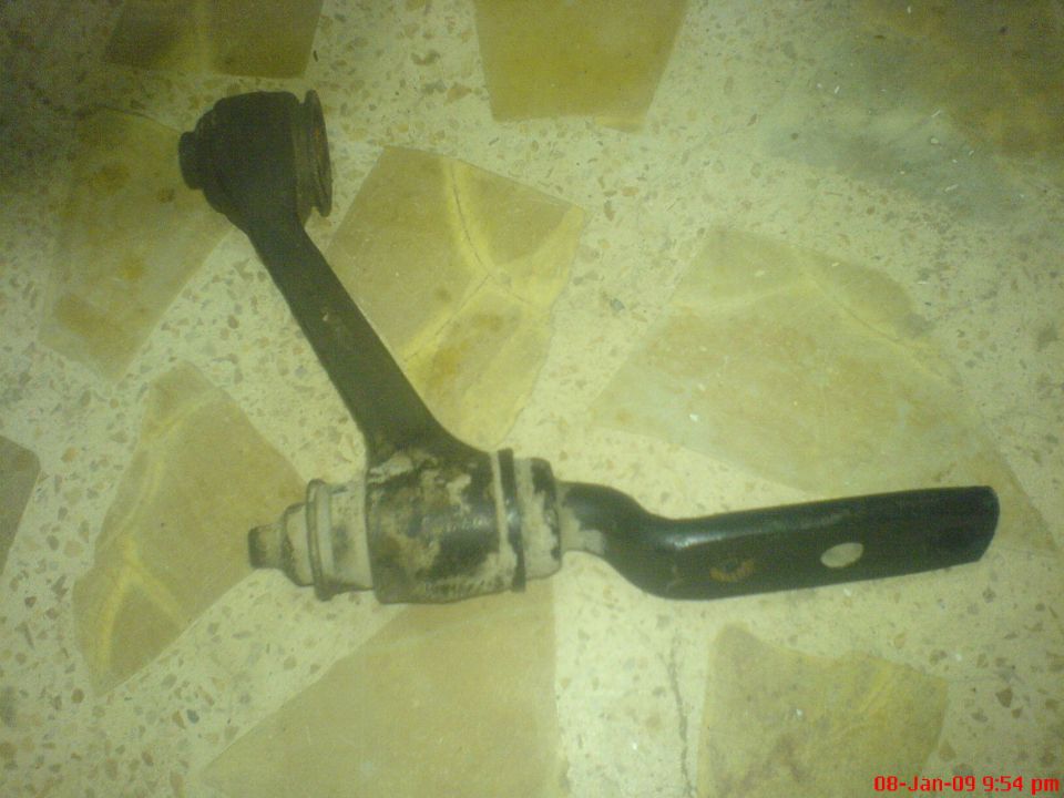 Black suspension control arm with rubber bushings on a speckled floor.