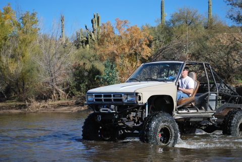 1986 Toyota Hilux - A modified beige Toyota pickup truck with a roll cage drives through shallow water in a desert environment with saguaro cacti and trees in…