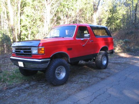 1989 Ford Bronco - A red 1989 Ford Bronco is parked on a dirt road next to a wooded area. The SUV has large off-road tires and a black bug deflector on the ho…