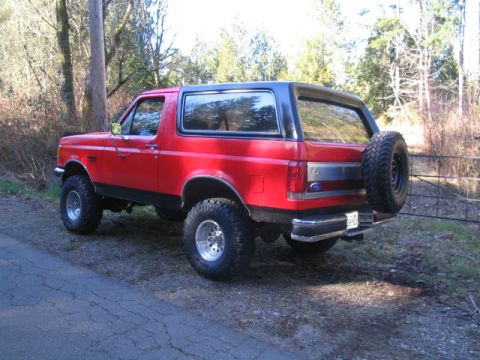 1989 Ford Bronco - The rear driver's side quarter view of a red and black 1989 Ford Bronco is parked on a gravel shoulder next to a paved road. A spare tire i…