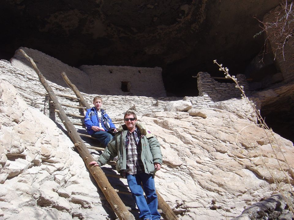 Two people on a wooden ladder in front of cliff dwellings.