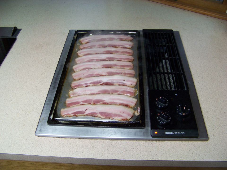 Raw bacon strips cooking on a Jenn-Air griddle.