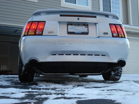 1998 Ford Taurus - The rear view of a silver Ford Mustang Cobra is shown parked on a driveway covered in patchy snow. The car features clear taillights and du…