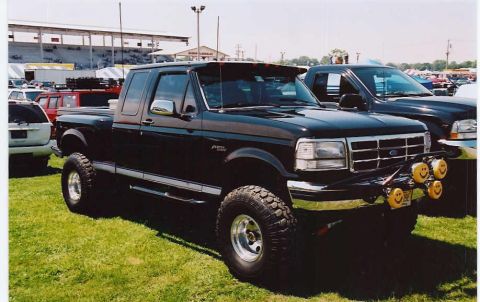 1994 Ford F-150 - A lifted black 1994 Ford F-150 pickup truck is parked on a grassy field next to another dark-colored truck.