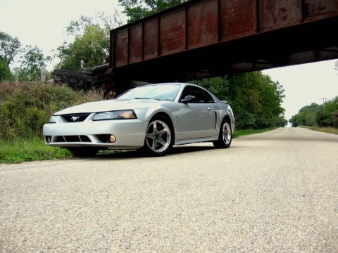 1998 Ford Taurus - A silver Ford Mustang coupe is parked on a gravel road under a rusty metal bridge. The car is viewed from a low angle, showing its front en…
